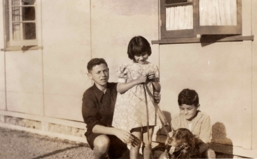Three 
   children in front of a distinctive Pahuatua hut. The older boy has his arm around his sister, who is standing in the 
   middle, head down, holding the lead, and the younger boy has his arms around the spaniel