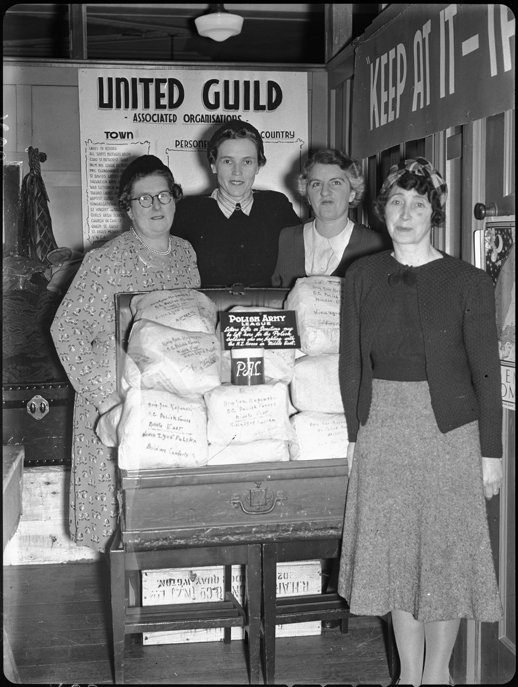 A black and photograph 
   of the four women, three behind a trunk full of packed goods, and Ann in front. The words United Guild Associated 
   Organisations are a backdrop.