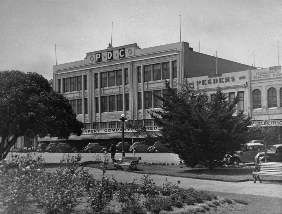 A black and photograph of 
   the front of the store, slightly obscured by trees in the Square. PDC clearly seen on the top facade. 1940s cars in front