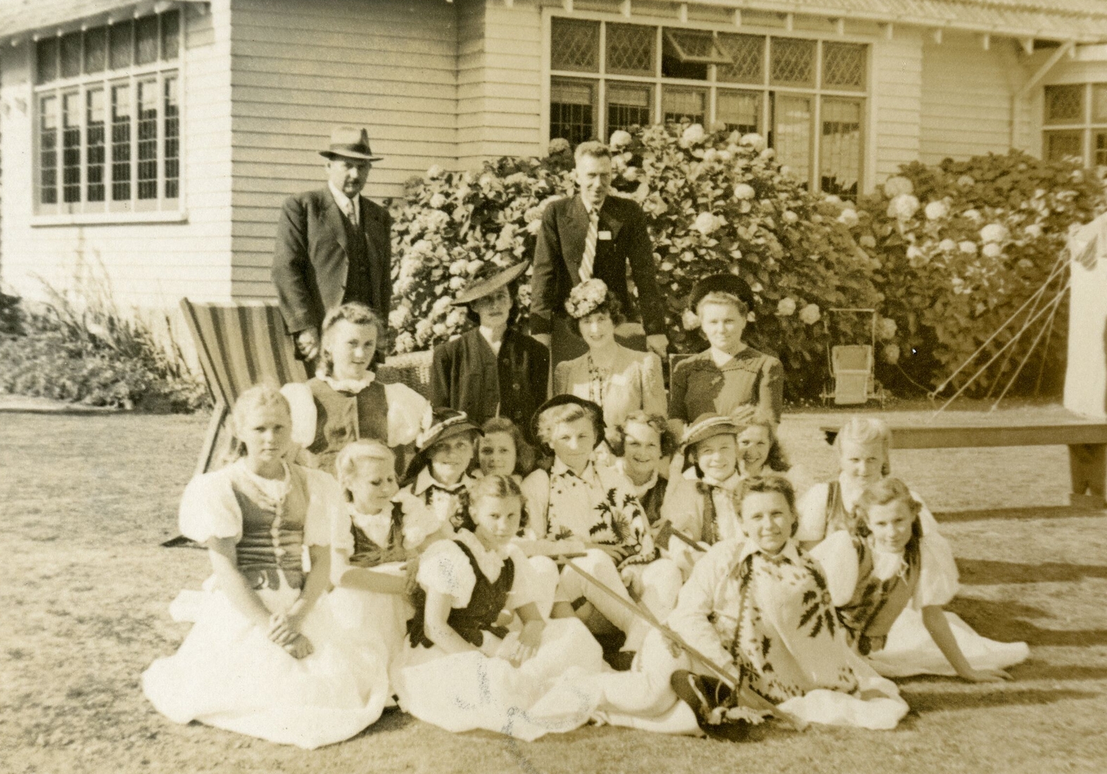 Sepia 
   shot of a group outside a house, two men standing in the back, three women sitting, and a group of girls in Polish 
   national costums in the front.