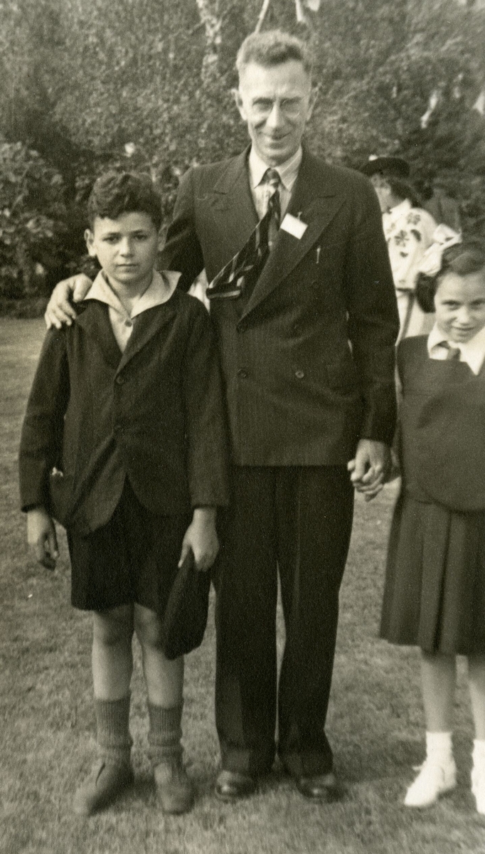 Standing, William smiling with his arm around Jan, and holding Henryka's hand, Henryka, head slightly down and 
   smiling slightly. Jan looks stoic. He is wearing a jacket with arms too short, buttoned incorrectly, and with a pocket 
   bulging. His collar is also up.