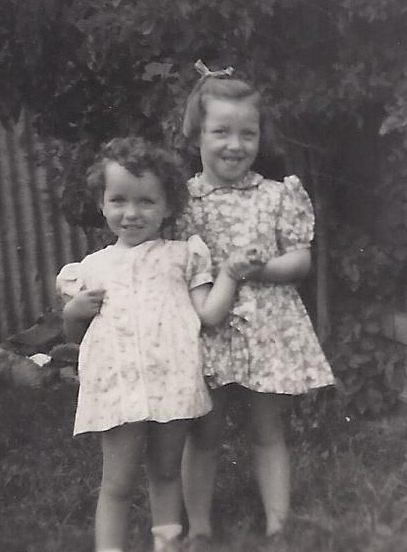 Faye and Carol in 
summer dresses, Faye behind Carol and holding her hand