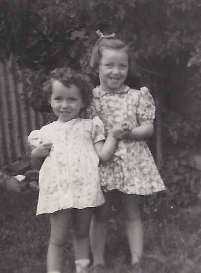 Faye and Carol in 
summer dresses, Faye behind Carol and holding her hand