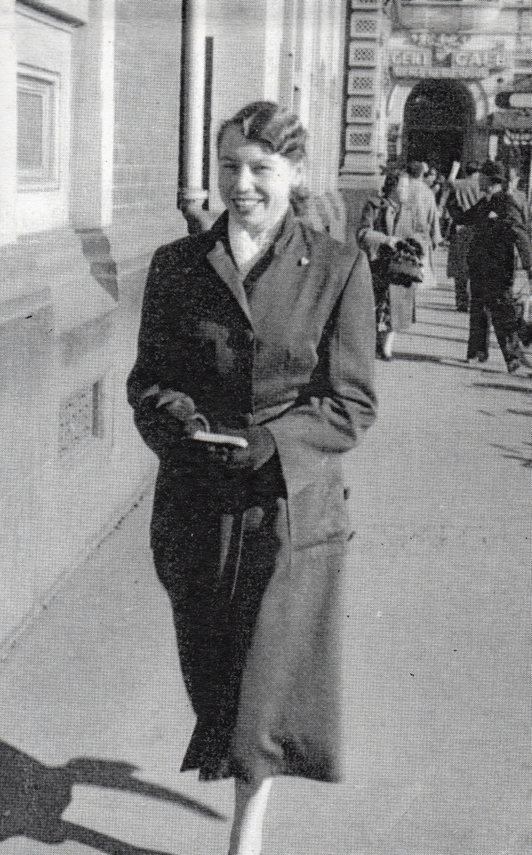 Henia striding  
along a street, smiling widely and wearing a skirt suit