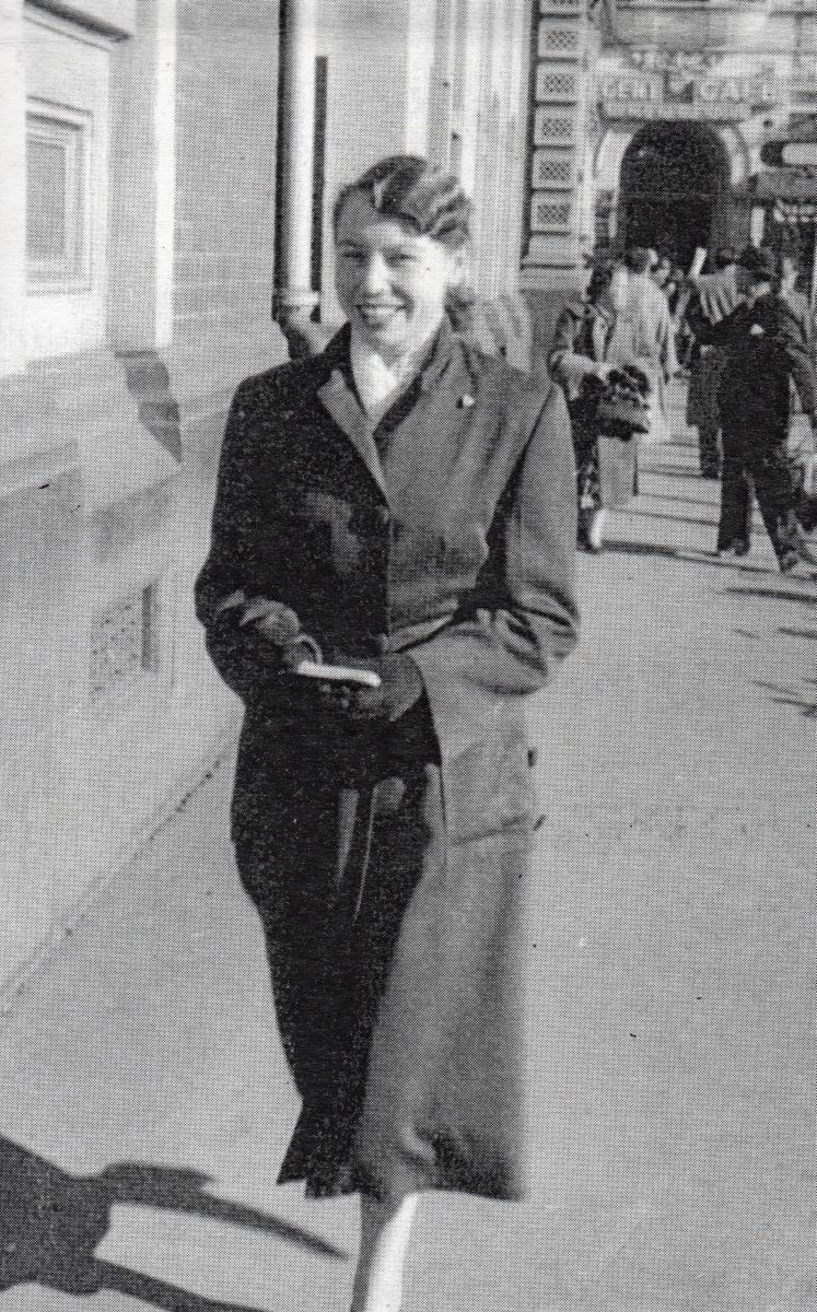 Henia striding  
along a street, smiling widely and wearing a skirt suit