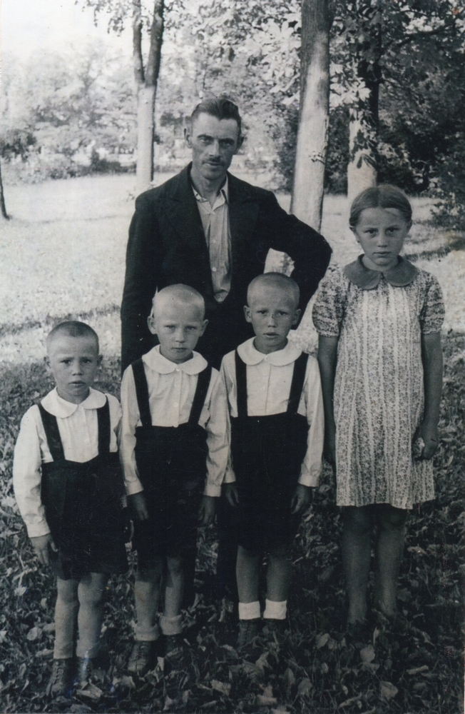 A photograph of Henryk standing with and four of his children, the boys dressed the same, in dark shorts and shirts and 
Irena in a dress. Serious children. 
