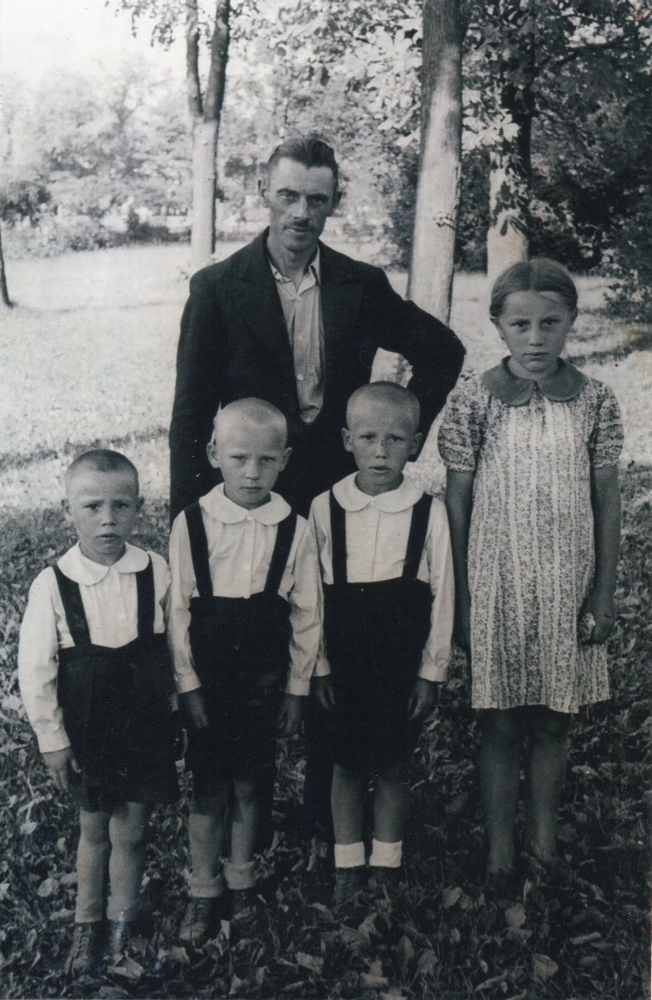 A photograph of Henryk standing with and four of his children, the boys dressed the same, in dark shorts and shirts and 
Irena in a dress. Serious children. 