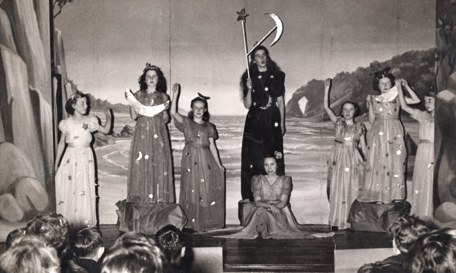 A scene on 
stage at Villa Maria. Seven girls in long dresses are standing at the back and Henia is sitting serenely middle stage