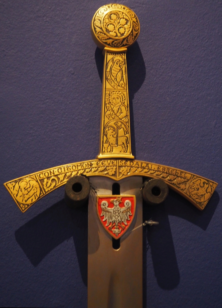 The hilt of the 
sword, and some of the blade, on a purple background. The handle is intricately decorated with a Letin inscription and 
symbols of the four Evangelists.