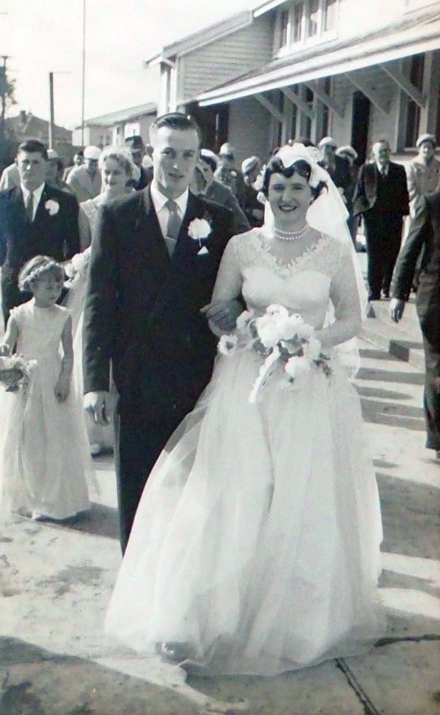 The newlyweds among their guests outside the school. The bridal party seems to have been stopped, because other 
   guests seem to be moving.