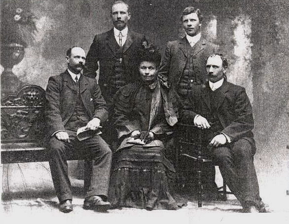 A black and 
white studio photograph of Franciszka sitting between two sons, and with the other two on either side behind her shoulders. 
They are all wearing formal attire. Franciszka has a black hat, holding black gloves, and has her eyes closed.