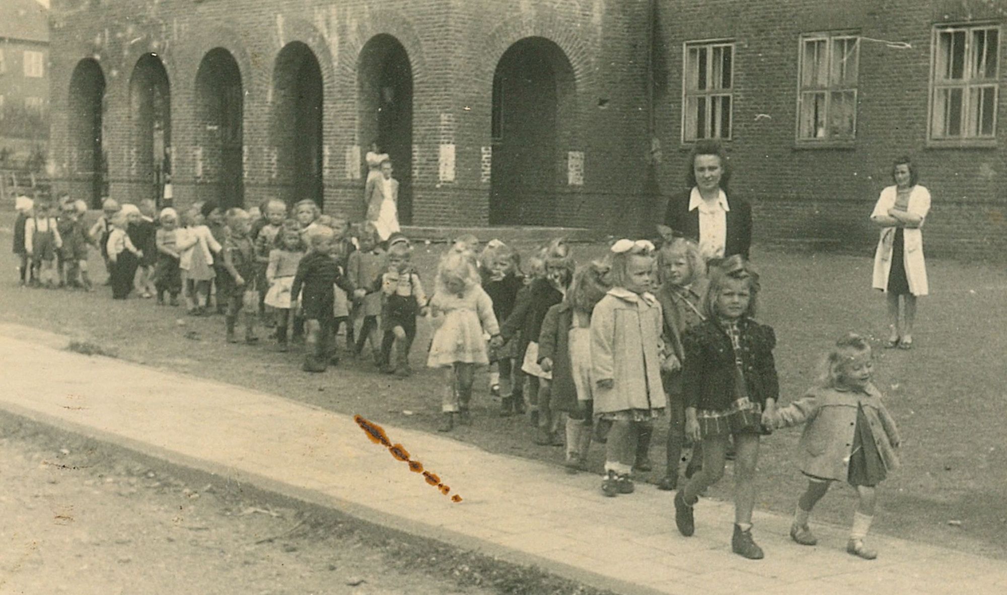 A crocodile of 
children walking from left to right along a path in front of the stark Flensburg DP buildings