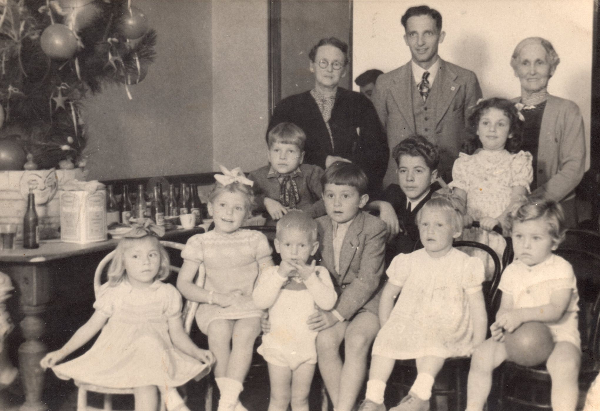 A group of 
refugees at a Christmas function, nine childen, five sitting in the front on chairs, and three adults standing behind them. 
Part of a Christmas tree is on a table to the left. 