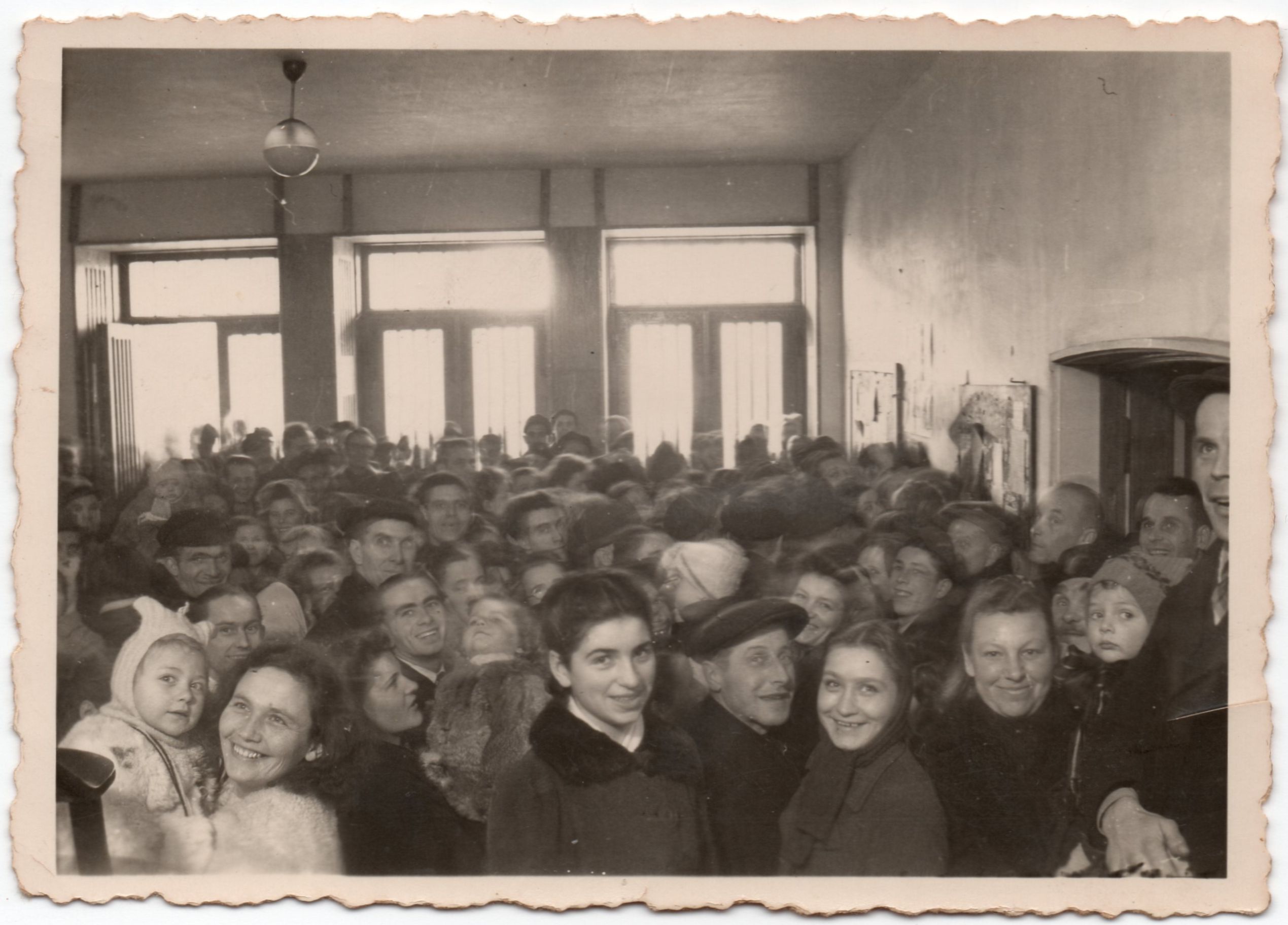 An 
immigration hall packed with new immigrants. People fill the bottom of the photograph. The top half shows a plain room with 
three large windows at the back.