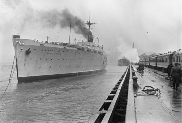 The ship alongside 
the wharf, which has a train waiting. The ship, and one behind, are spouting smoke. A man is walking front right away from 
the camera