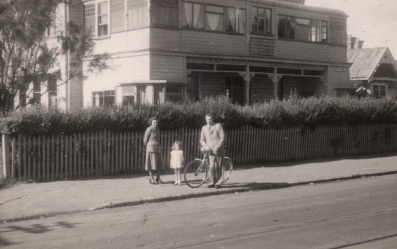 The Kuźmiuks are 
standing on the pavement outside the hotel, which looks like a large house, and which borders the pavement with a low fence 
and a hedge taller than Pawel, who is astride a bicycle.