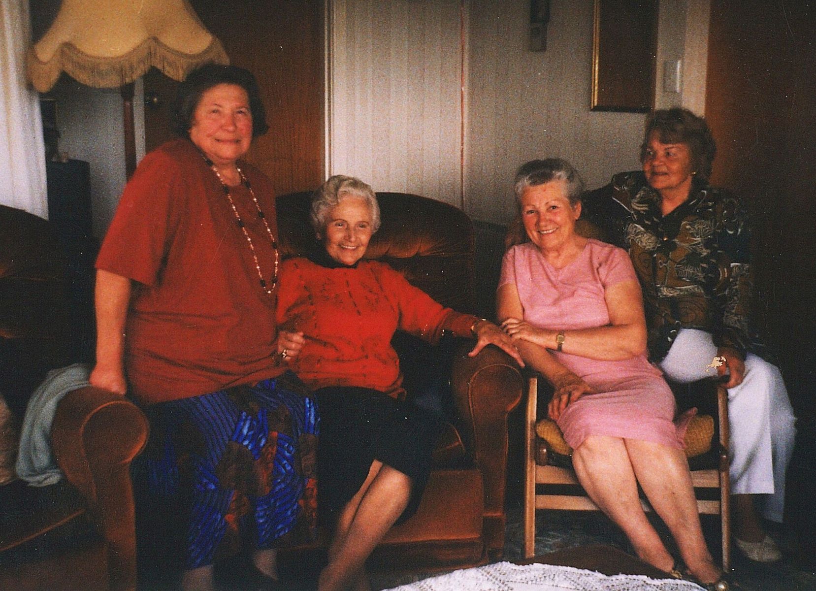 Four 
middle-aged women in a lounge, the middle two sitting on chairs and the outer two perched on the armrests. All are smiling.