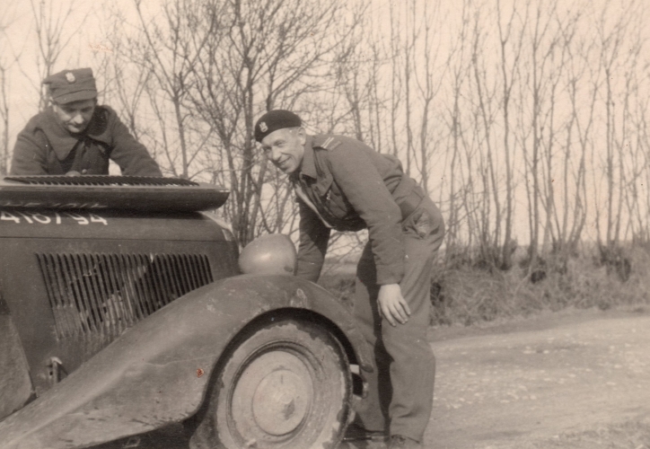 Paweł  Kuźmiuk 
looks up and smiles as he leans over the open engine of an army vehicle. Another soldier is head down over the engine. Flat 
fields, and leafless thin trees line the road behind him.