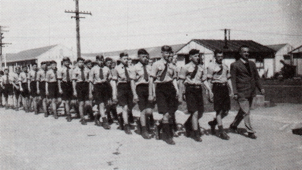 black and white pic 
   of a group of boys marching along a road, back ground is barrack-like buildings