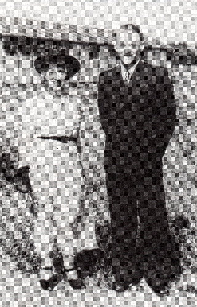 black and 
white pic of the two in a field with a barrack in the background