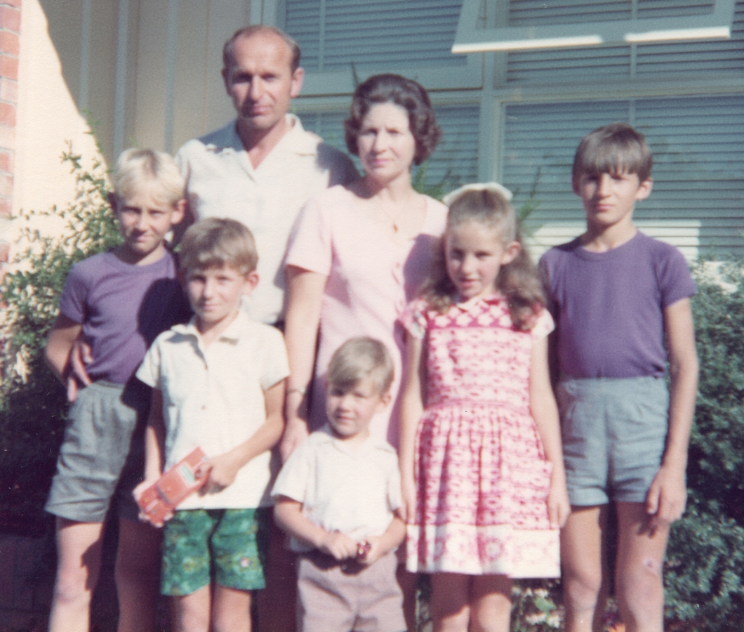 A coloured but 
slightly blurry summer photograph of the family outside a house. The parents are at the back, the children in front of them. 
the two boys on either side are wearing matching purple tee-shirts and grey shorts. 