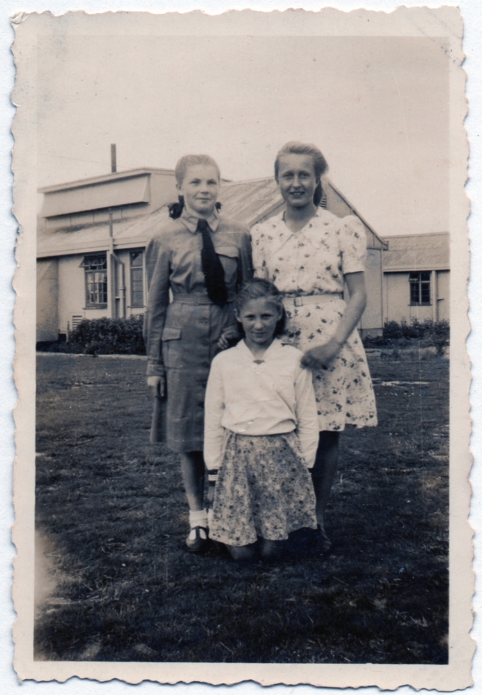 A sepia photograph of 
three girls, camp building in the background. Zofia in uniform has her hair neatly tied back and is standing very upright 
with her arm next to her side. Next to her is a girl about her age in a short-sleeved flowered dress. Irena is kneeling in 
front of them. She is wearing a flowered skirt and a white long-sleeved shirt.