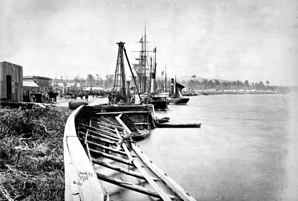 Black and white 
photograph of the wharf on the left, with several vessels with masts alongside, and buildings in the harbour's curve in the 
distance.