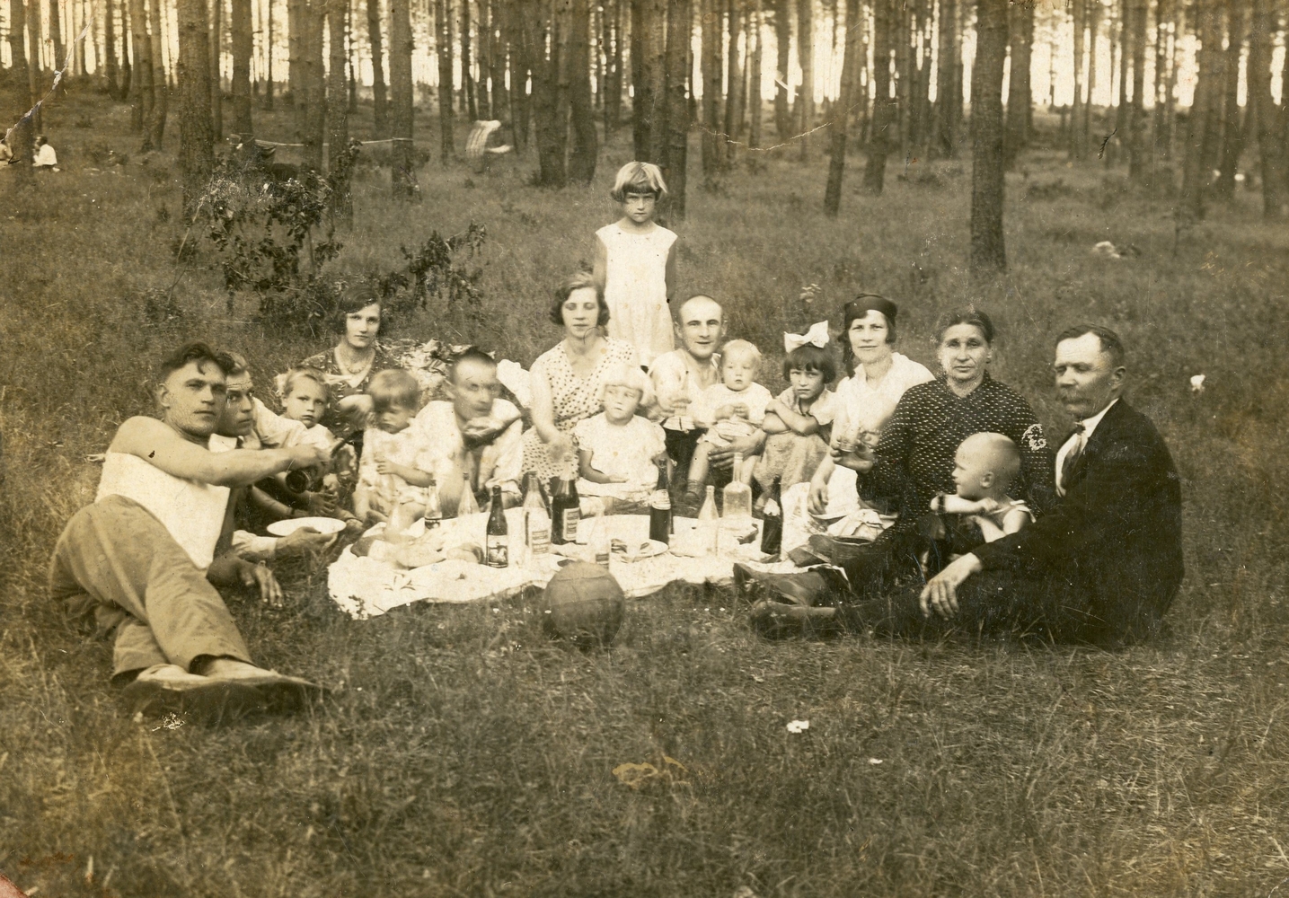A sepia 
photograph of 15 people of three generations sitting around a tablecloth in a forest clearing.