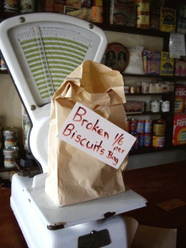 Paper bag on an 
old-fashioned scale with the words Broken biscuits 1/6 per bag