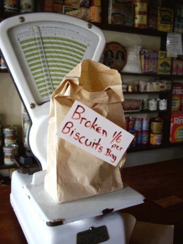 Paper bag on an 
old-fashioned scale with the words Broken biscuits 1/6 per bag