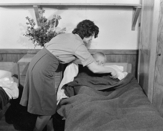 Young Polish boy 
being tucked into bed by unidentified woman, in skirt and shirt, possibly WAAF uniform