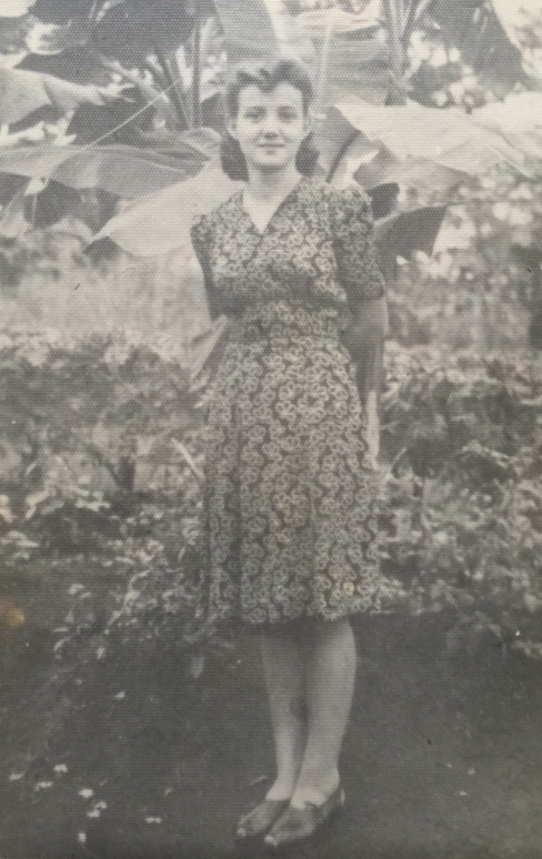 Joanna in 
a dress, hands behind her back, standing in a garden.