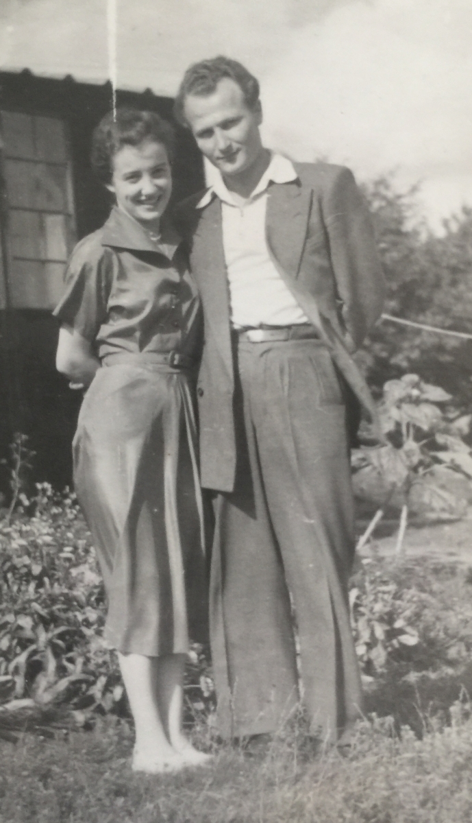 Joanna and 
Tadeusz standing in a garden in front of a house, she in a light, collared dress and he in a light suit and open-necked 
shirt.