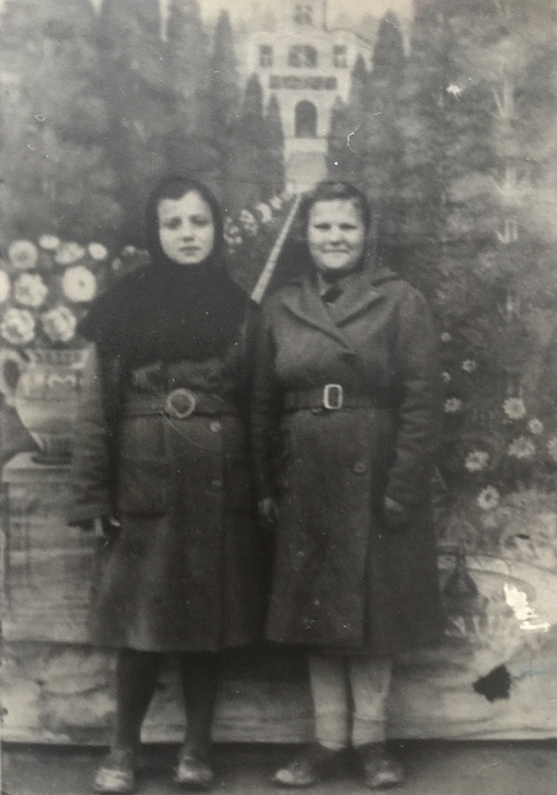 Joanna 
and a friend, dressed in coats, pose in front front of a studio backdrop of a tree-lined vista with a palace in the 
background.