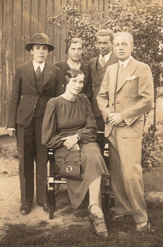 Bronisława 
Meder sitting on a chair with four people standing behind her, Józef to the front right and her sister between the other two 
men; photograph taken in a garden.