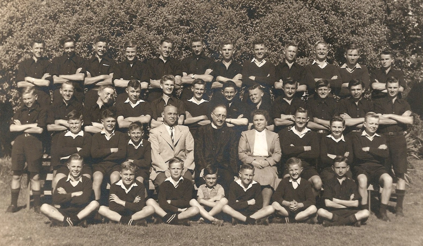 More formal group 
photo, boys dressed in dark uniforms, all arms folded. Mr and Mrs Tietze in light clothes, priest sitting between them.