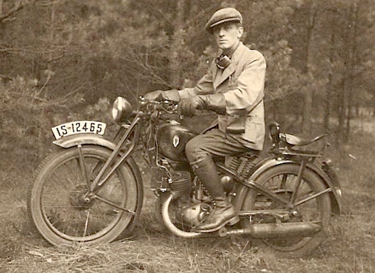 Josef sitting 
on a motorbike, a wooded background. He is wearing a cap and light jacket, and has goggles around his neck and a black 
rectangular camera bag attached to a strap that seems to hang from the unseen shoulder.