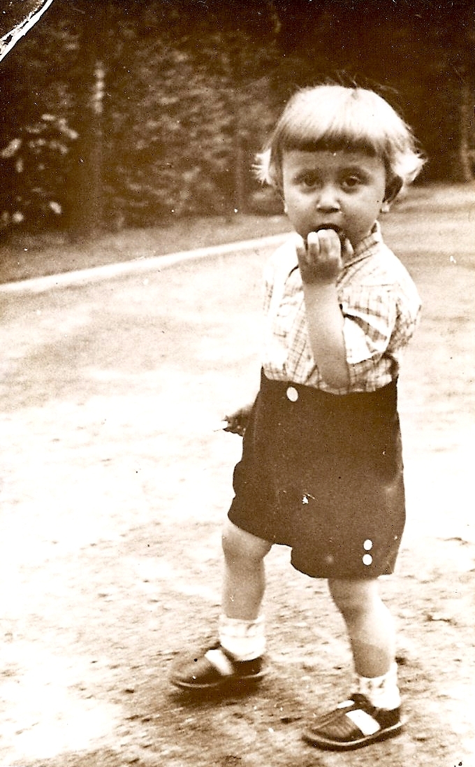 Blond Kazik 
dressed in short-sleeved shirt and shorts, socks and sandals, hand in mouth but looking at the camera.