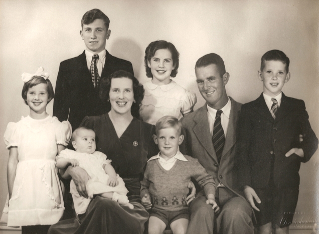 A studio photo 
of Kazik and the Campbell family. Mr and Mrs Campbell and their three oldest children are all smiling widely: Kazik less so. 
Baby on Mrs Campbell's lap and the second-youngest between his parents. Photo exudes positivity.
