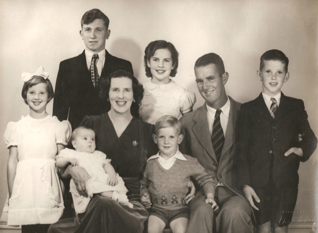 A studio photo 
of Kazik and the Campbell family. Mr and Mrs Campbell and their three oldest children are all smiling widely: Kazik less so. 
Baby on Mrs Campbell's lap and the second-youngest between his parents. Photo exudes positivity.