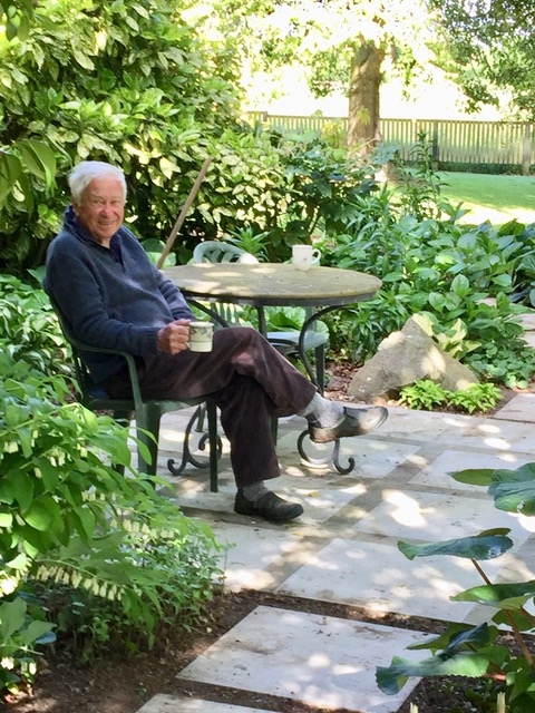 Kazik sitting at a 
table in the garden with sloppy jumper, slippers and a mug in his hand; smiles at the camera.