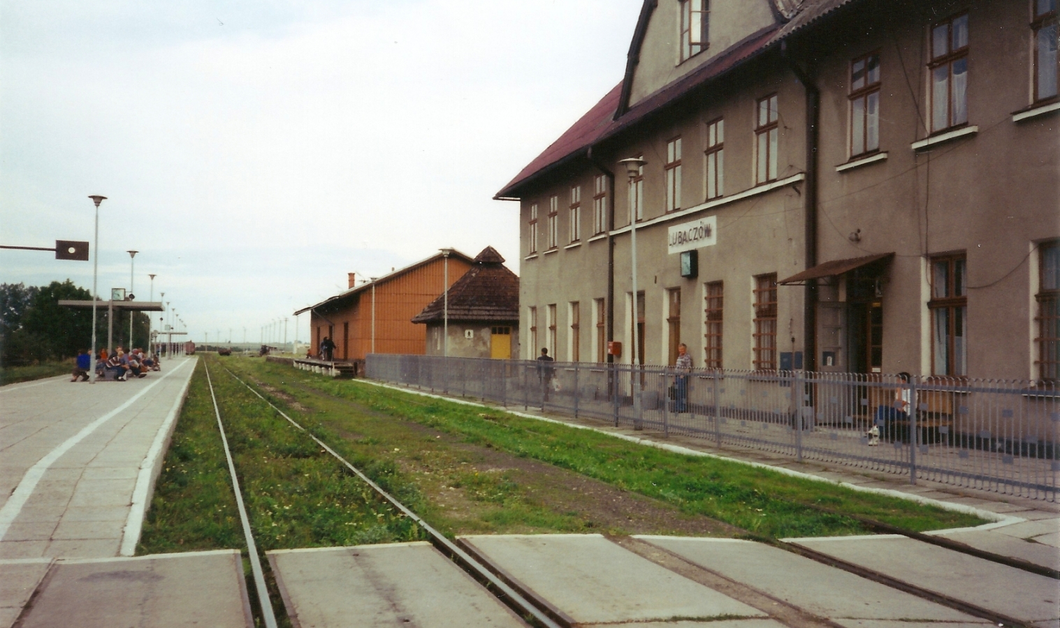 The outside 
of Lubaczów railway station. Kazik stands under the station sign, small with the whole image.