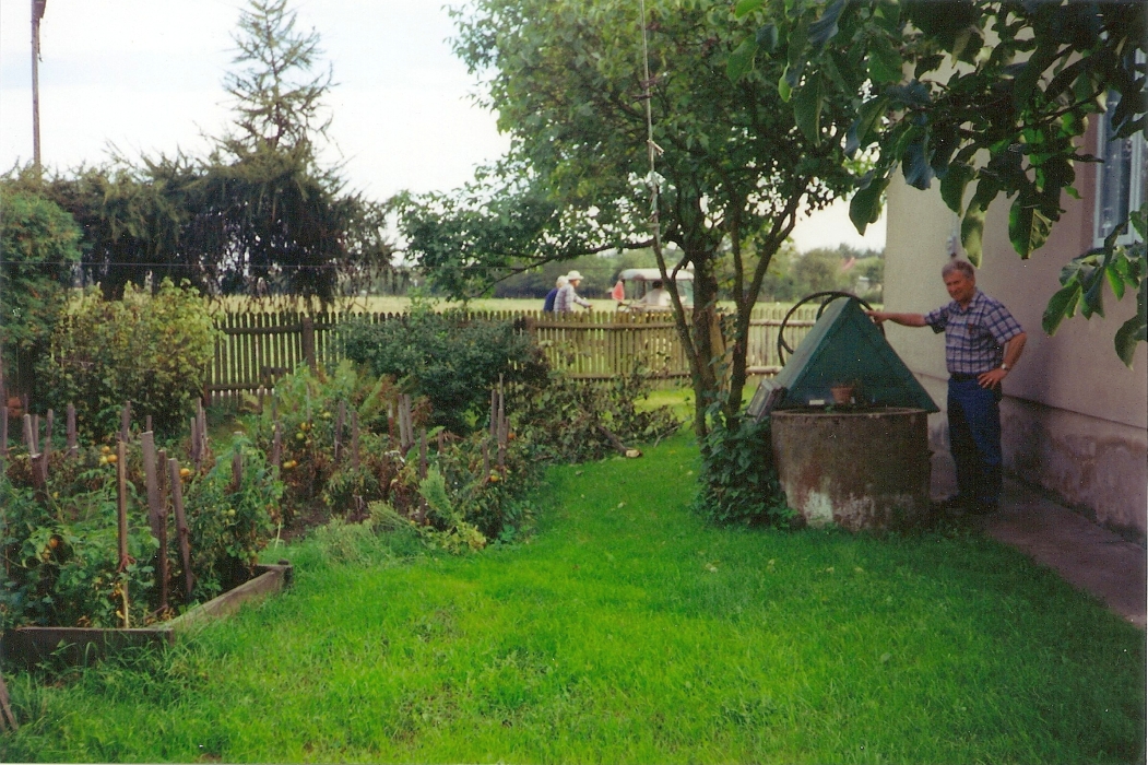 Kazik next to 
the well between the tree and the house, low wooden picket fence and people walking past, and ripening tomatoes and other 
vegetables on the other sunny side.