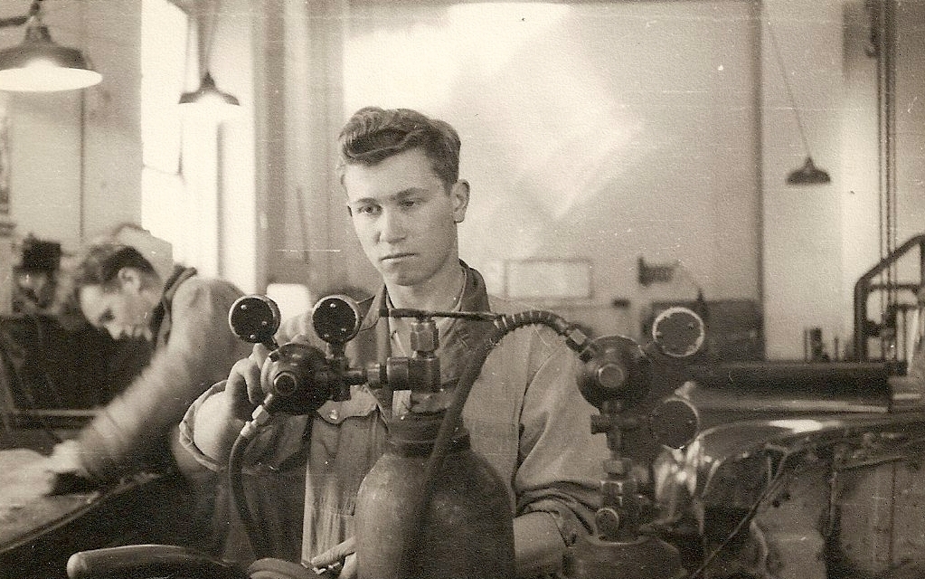 Kazik stands 
behind a gas cylinder as he looks at, and has his hand on, the attached gas regulator. One other apprentice in background.