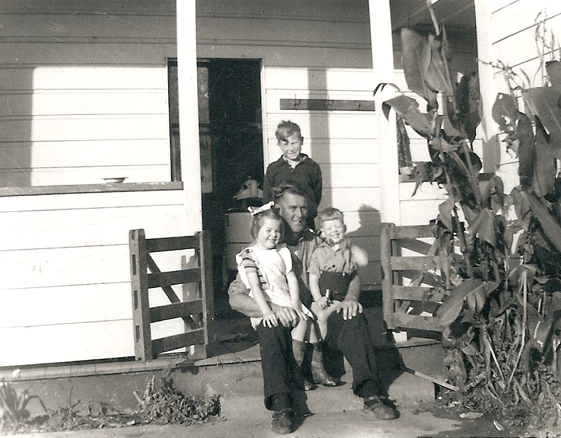 Outside a white 
weatherboard door, on a porch, Kazik stands behind John Campbell, who is sitting on the step with his two oldest children on 
His knees.