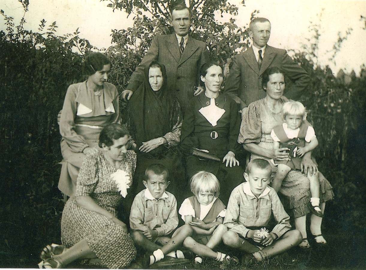 Maria Meder, severe 
and dressed in black with a black scarf over her head, sits with her daughters, and grandchildren. Sons-in-law stand at back.
Taken in a garden.