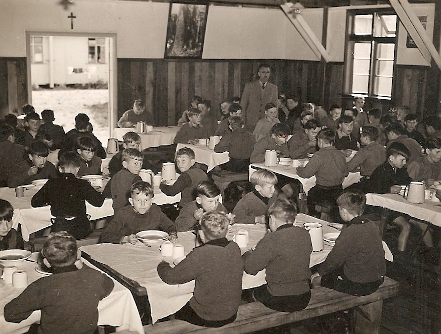 The inside of the 
dining room, four rows of tables with bench-seats, white tablecloths and boys eating. A man is at the back.