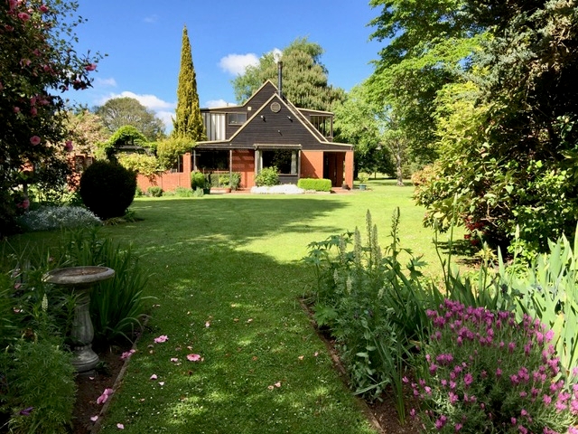 A wide shot from a grassed 
path towards the house, pink Camellia flowers on path, pink perennials on other side, clipped hedging, many trees.