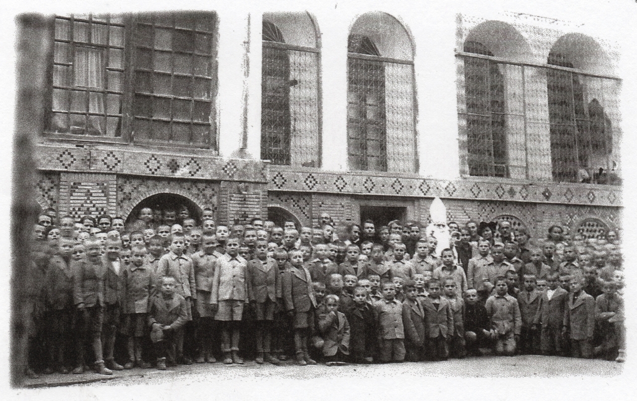 A far more sombre 
black and white group photograph of the boys, wearing long-sleeved jackets, in front of their building, ornately decorated. 
Above them are the arch-shaped dormitory windows