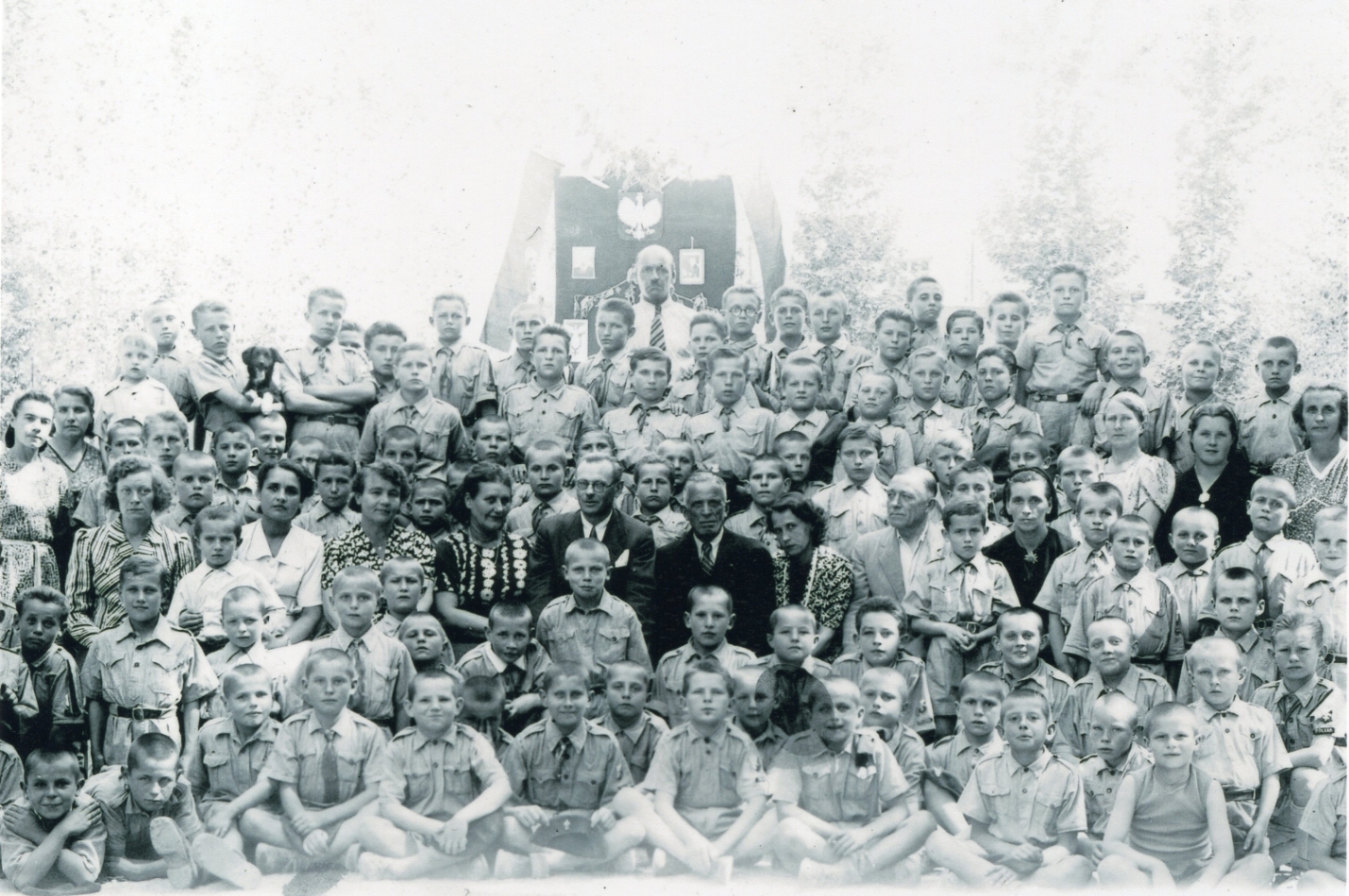 A black and white 
group photograph of at least 60 young boys, wearing mostly military-style uniforms, and caregivers, in front of a banner with 
a white eagle. Few smiles, but one boy holding a black dog.